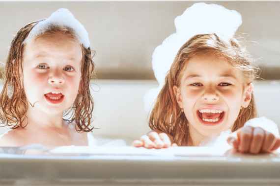 Zwei kleine Mädchen sitzen in einer Badewanne, haben Badeschaum am Kopf und lachen herzhaft