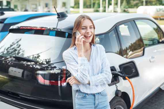 Transportation. Young woman on electric car having stop at charging station leaning on vehicle talking on smartphone laughing cheerful while charging