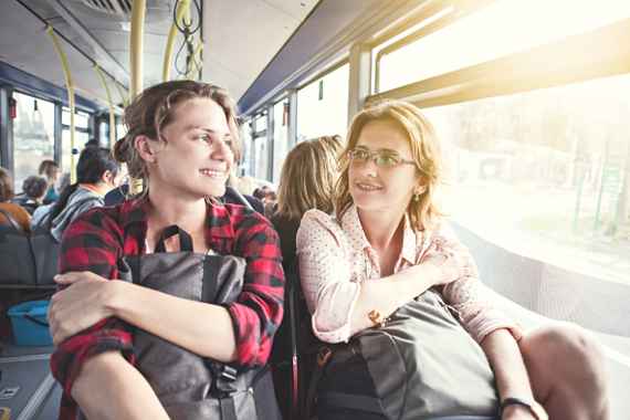 Zwei junge Frauen sitzen im Bus mit ihren Rucksäcken und sehen sich an.