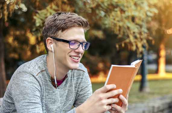  Junger Mann mit Brille sitzt in einem Park, liest und hört mit Kopfhörern Musik.