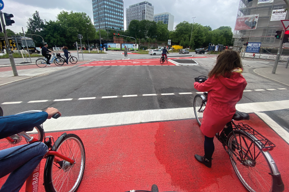 Radfahrende stehen an einer ampelgeregelten Kreuzung, die für Radfahrende rote Bodenmarkierungen aufweisen