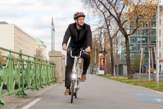 Ein Mann im Anzug und mit Helm fährt auf einem Faltrad auf einem gemischten Geh- und Radweg.