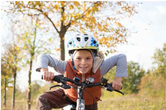 Bub mit Fahrradhelm stützt sich am Lenker seines Fahrrades ab und lächelt dir fröhlich ins Gesicht