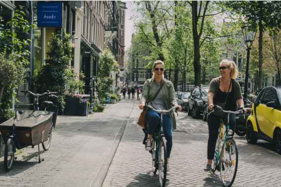 zwei Damen auf dem Fahrrad auf der Straße in einer kleinen Stadt mit viel Blumen am Gehsteig