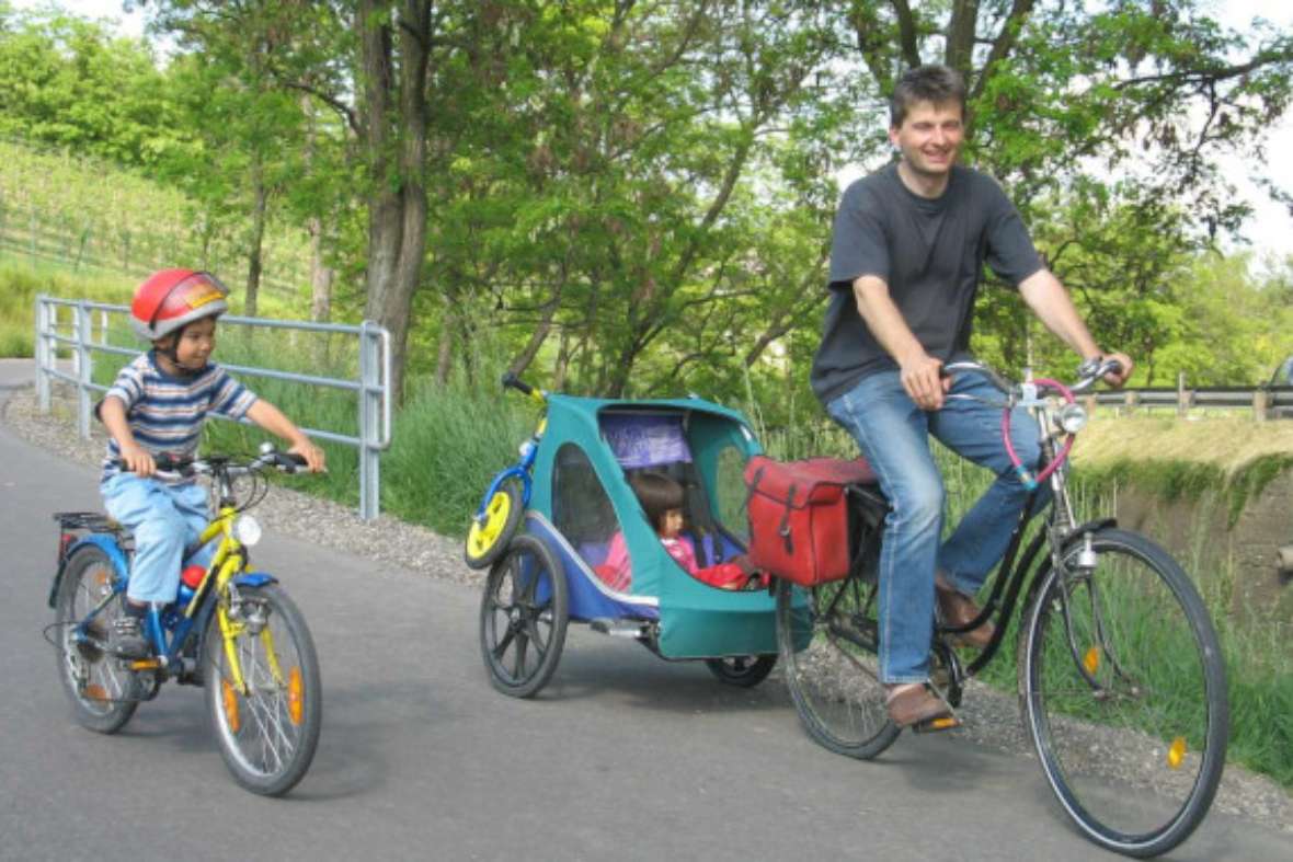 Kind mit Helm auf Fahrrad und Erwachsener mit Fahrradanhänger