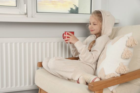 Little girl with cup of hot drink near heating radiator indoors