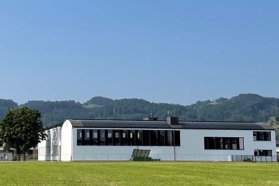 Ansicht der Volksschule Höchst Kirchdorf, im Vordergrund ein Sportplatz, links im Hintergrund eine Kirche, dahinter bewaldete Hügel