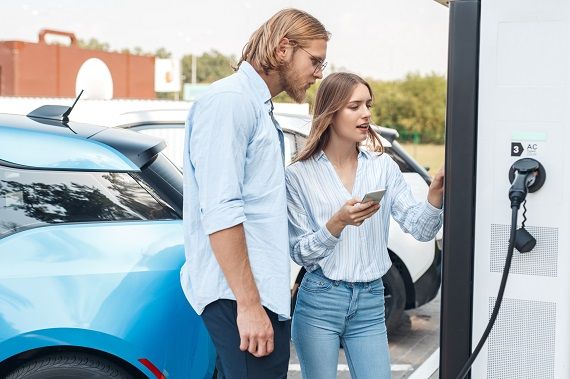 Traveling. Young couple traveling by electric car stopping at charging station girlfriend with smartphone choosing voltage on screen pensive together with boyfriend