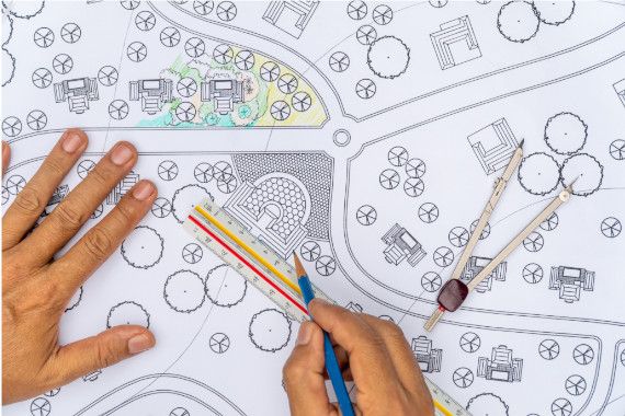 Skizze eines Bebauungsplan mit vielen Bäumen und wenigen Straßen, eine Hand hält einen Bleistift, Zirkel liegt am Plan