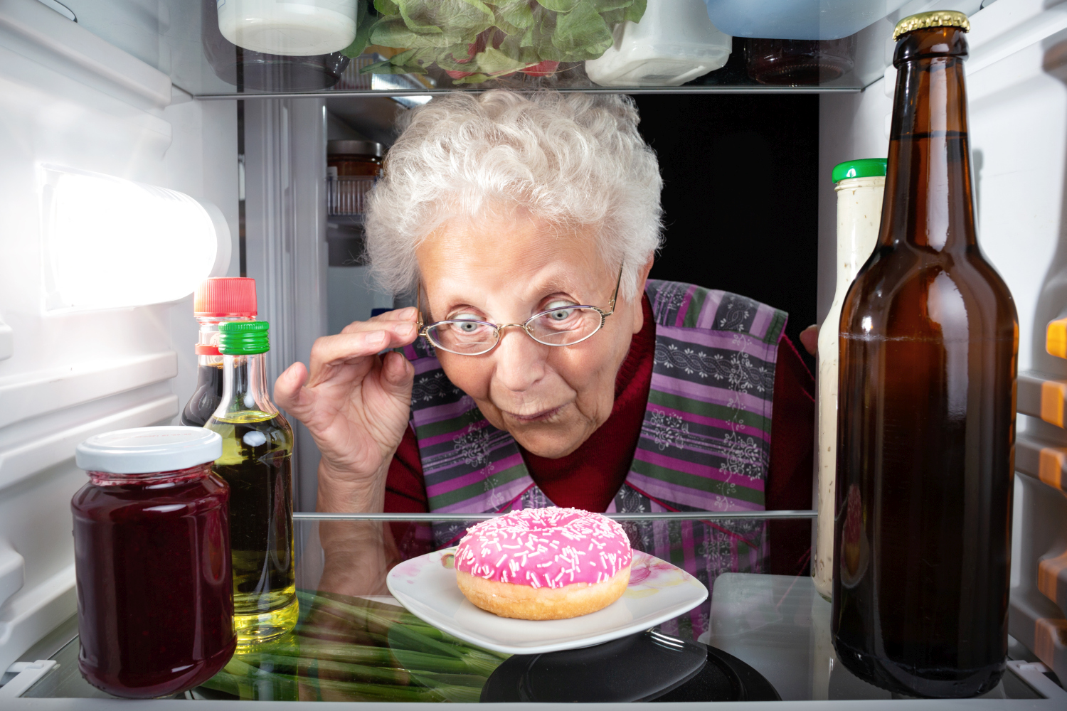 Eine alte Frau steht erstaunt an der Kühlschranktür und schaut erstaunt auf einen pinken Donut