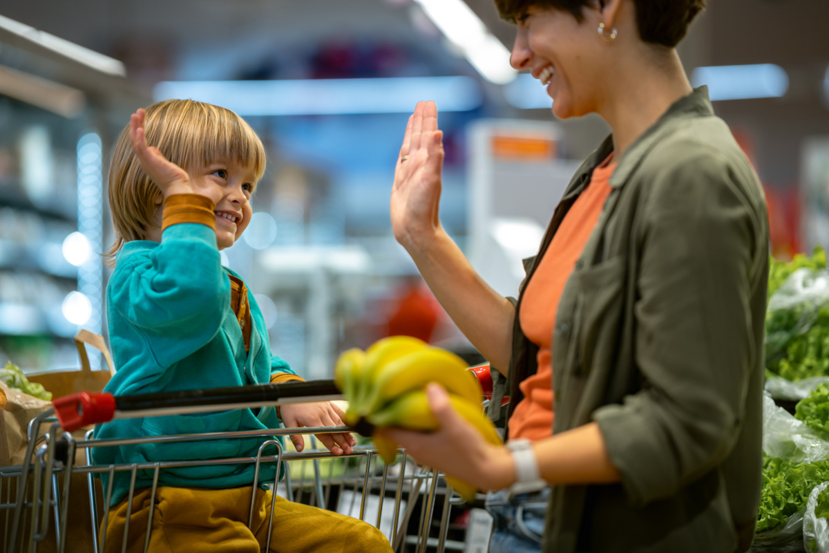 Kind im sitzt im Einkaufswagen und Mutter mit Paprika in der Hand steht daneben. Sie schauen sich in die Augen und schlagen High-Five