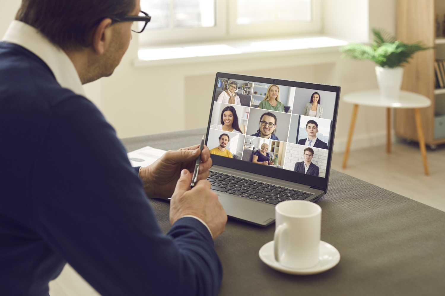 Mann mit Brille sitzt vor Laptop und hält Videokonferenz. Am Bildschirm verschiedene Gesichter