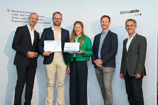 Menschen mit einer Urkunde in der Hand vor einer Fotowand. Sie lächeln in die Kamera.