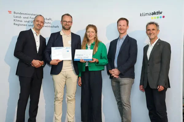 Menschen mit einer Urkunde in der Hand vor einer Fotowand. Sie lächeln in die Kamera.