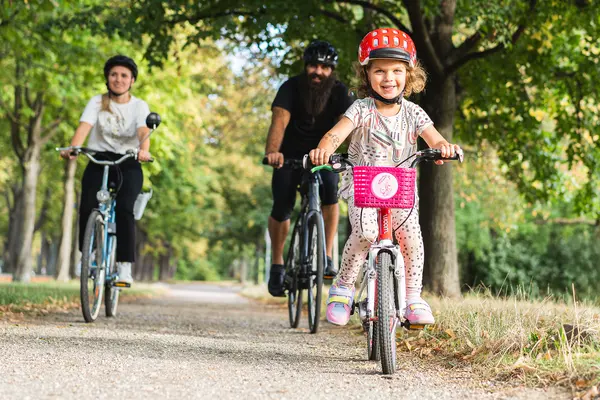 Ein Kind auf einem Kinderfahrrad und mit Helm radelt durch einen Park. Im Hintergrund radeln die Eltern hinterher.