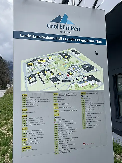 Ein Wegweiser der „tirol kliniken“ in Hall in Tirol mit Informationen zum „Landeskrankenhaus Hall“ und zur „Landes-Pflegeklinik Tirol“. Der Wegweiser zeigt eine detaillierte Karte des Krankenhausgeländes mit farblich gekennzeichneten Gebäuden (Haus 1 bis Haus 6, Haus West und Haus E) sowie einer Liste der jeweiligen Stationen und medizinischen Einrichtungen.