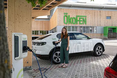 Elektrisches AUto steht unter Carport, Frau steckt Ladekabel an