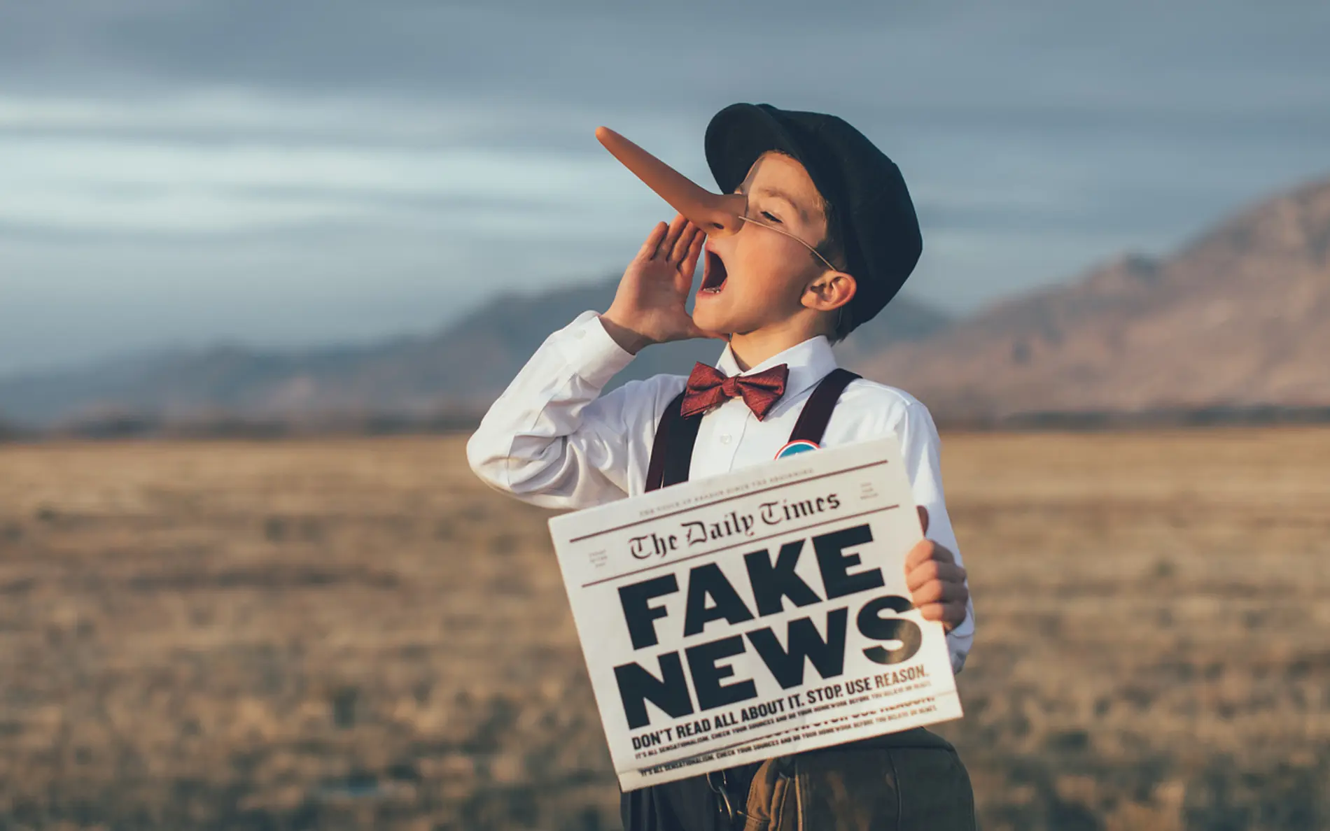 Ein Junge hält eine Zeitung in der Hand, auf der fett gedruckt Fake News steht und um seine Nase hat er eine Pinocchio-Nasenattrappe befestigt, die auf die Falschaussagen aufmerksam machen soll 