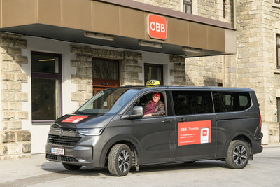 Ein dunkelgrauer Shuttlebus mit „ÖBB Transfer“-Aufkleber steht vor einem ÖBB-Bahnhofsgebäude. Am Steuer sitzt ein freundlicher Fahrer mit roter Kappe und lächelt in die Kamera. Auf dem Dach des Fahrzeugs ist ein Taxi-Schild angebracht. Der Eingangsbereich des Bahnhofs mit ÖBB-Logo ist im Hintergrund sichtbar.