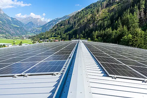 Umfangreiche Solarpaneele auf einem geneigten Metalldach, mit grünen Hügeln und Bergen im Hintergrund.