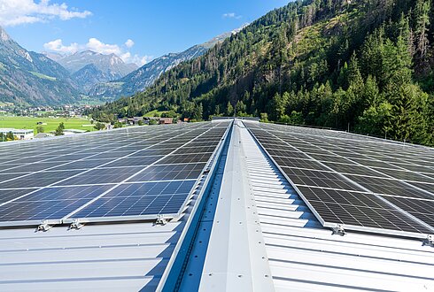 Umfangreiche Solarpaneele auf einem geneigten Metalldach, mit grünen Hügeln und Bergen im Hintergrund.