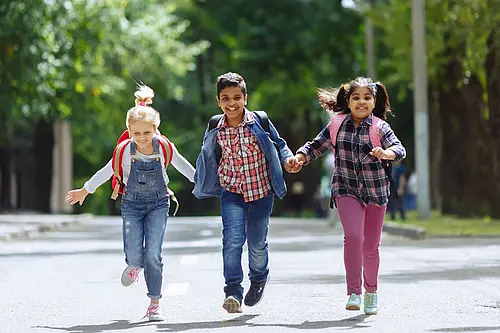 Gruppe von drei Kindern laufen Hand in Hand einen Weg entlang und lachen dabei. Sie tragen Schultaschen