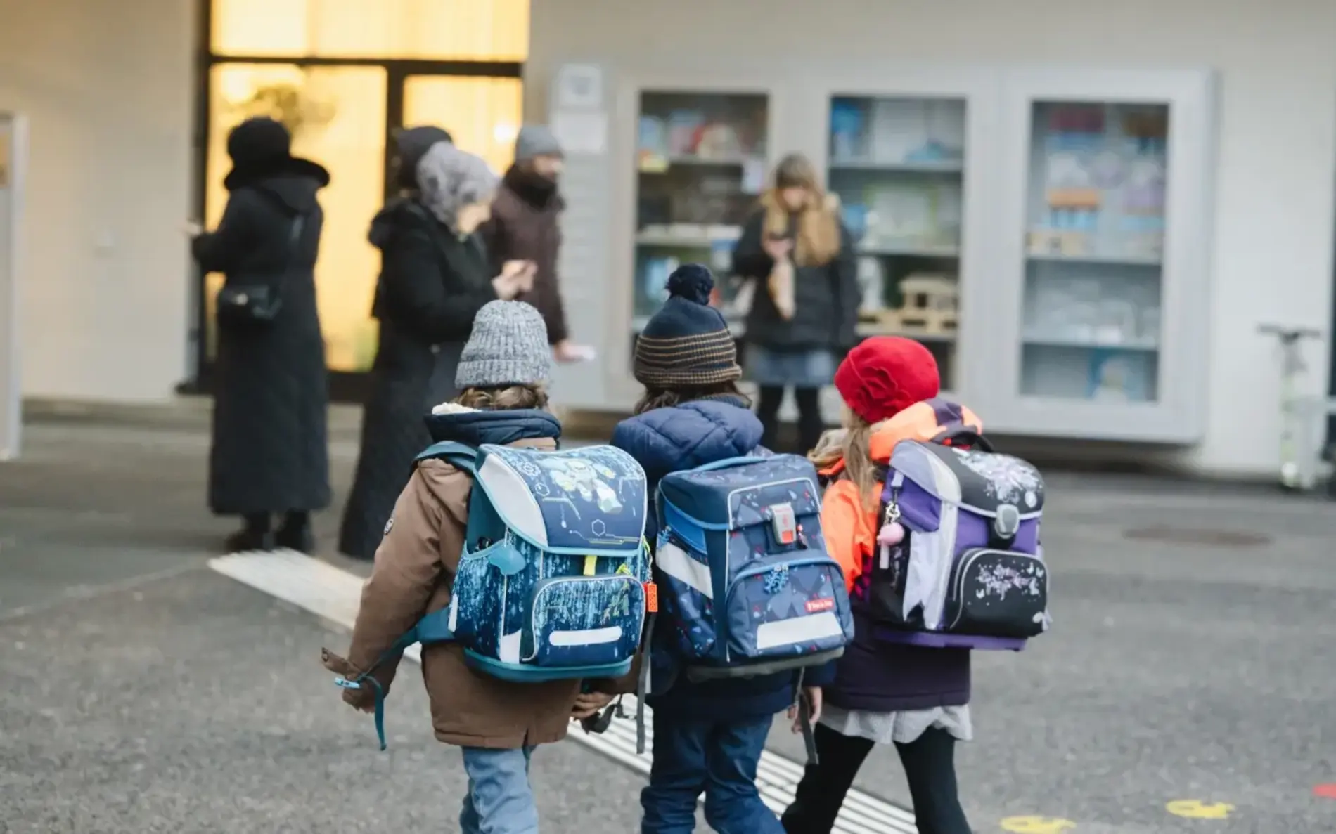 Drei Kinder mit Schultasche gehen über den Schulvorplatz, im Hintergrund wartende Eltern und eine Gebäudewand