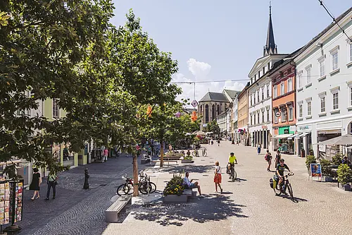 Breite Ansicht über den Hauptplatz Villach mit vielen Radfahrern und Passantinnen. Links viele Bäume