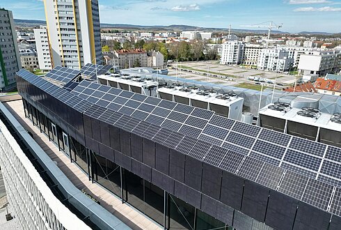 Blick auf ein Gebäudedach mit zahlreichen Solarpaneelen, im Hintergrund eine städtische Umgebung.