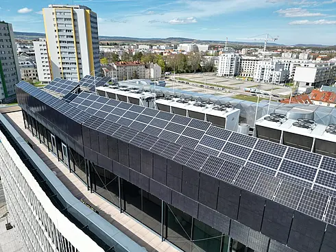 Blick auf ein Gebäudedach mit zahlreichen Solarpaneelen, im Hintergrund eine städtische Umgebung.