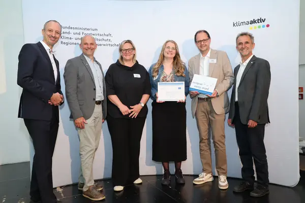 Menschen mit einer Urkunde in der Hand vor einer Fotowand. Sie lächeln in die Kamera.