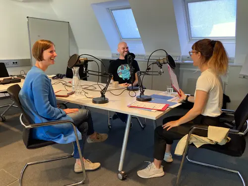 Johannes Naimer-Stach und Marion Zöchbauer sitzen mit der Podcast-Moderatorin Anna Breuer um einen Tisch. Sie lachen einander an. Auf dem Tisch stehen drei Mikrofone auf Stativen.