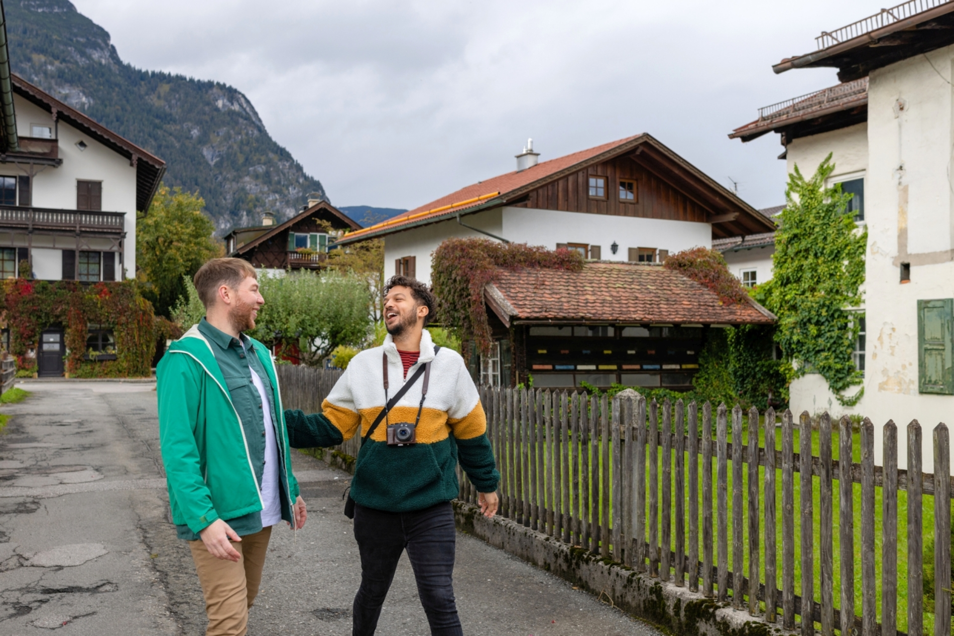 Zwei junge Männer spazieren durch ein typisch österreichisches Dorf. Im Hintergrund zwei ländliche Häuser mit begrünten Balkonen und ein Bergpanorama.