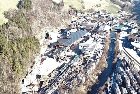 Luftbild eines Holzlagers mit Holzstapeln, Gebäuden, angrenzendem Waldgebiet und nahegelegenem Dorf.