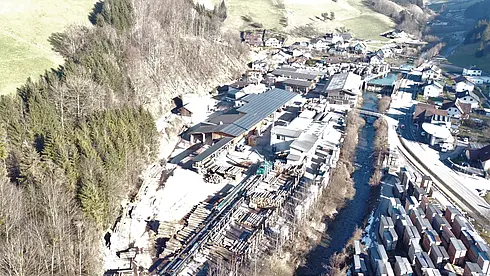Luftbild eines Holzlagers mit Holzstapeln, Gebäuden, angrenzendem Waldgebiet und nahegelegenem Dorf.