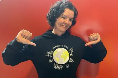 Karin Mühlsteiger zeigt mit beiden Daumen auf ihren Sweater. Dort zu sehen ist ein Run around the world Logo