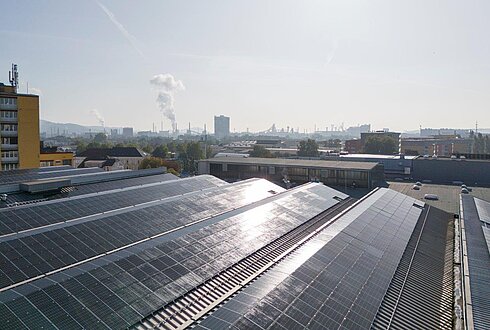 Photovoltaikanlage auf mehreren Dachflächen in einem Industriegebiet, im Hintergrund Stadtgebäude und Schornsteine.