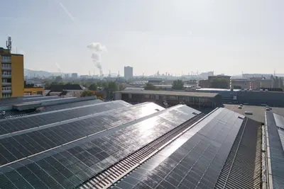 Photovoltaikanlage auf mehreren Dachflächen in einem Industriegebiet, im Hintergrund Stadtgebäude und Schornsteine.