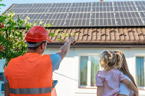 Ein PV-Installateur mit Warnweste und Helm zeigt in Richtung eines PV Daches mit PV-Paneelen. Daneben steht 