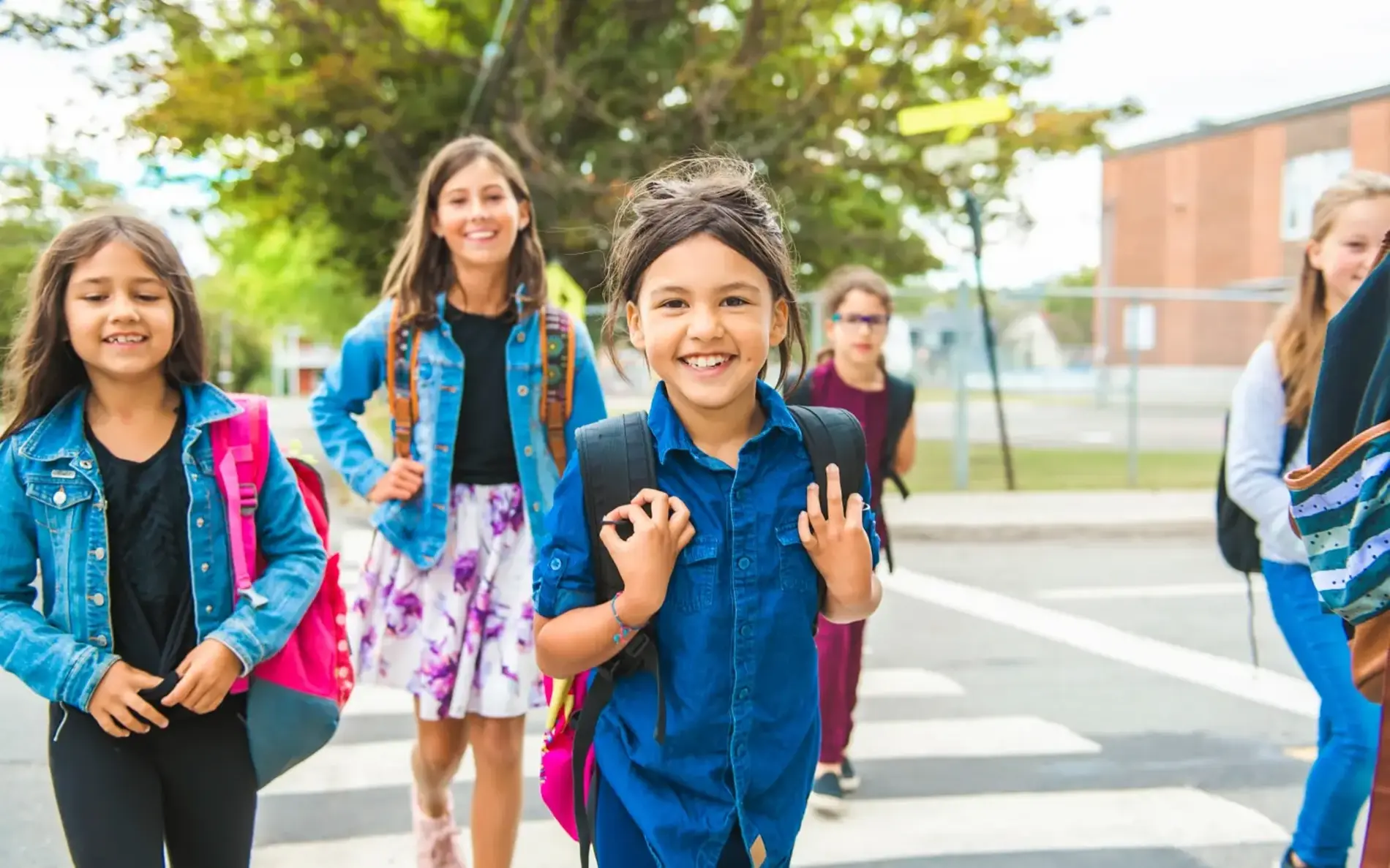 Kinder mit Schultasche gehen auf die Kamera zu - im Hintergrund ein Baum