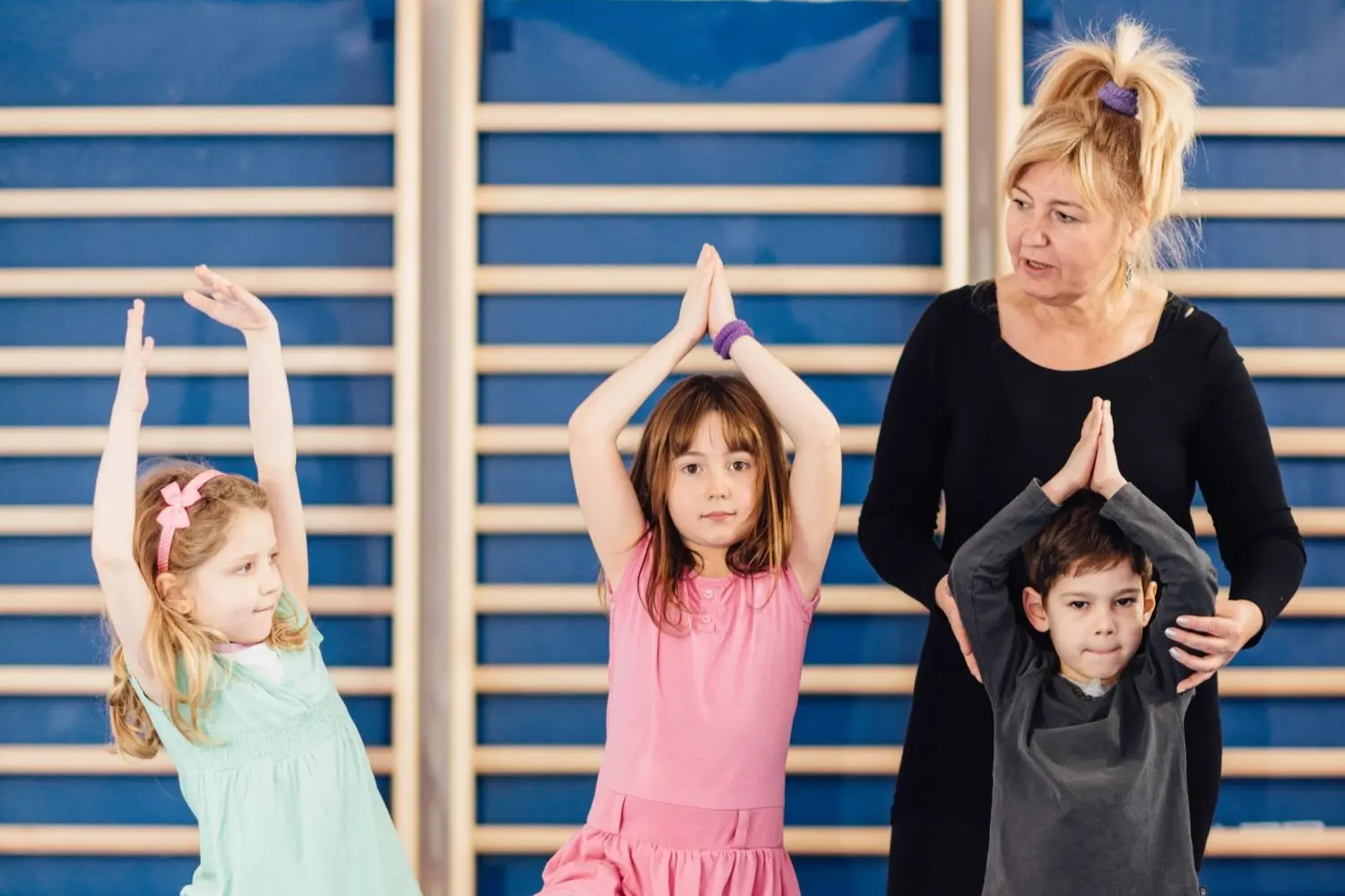 Drei Kinder und eine Lehrerin stehen vor einer Sprossenwand im Turnsaal. Die Kinder halten die Hände gefaltet über den Kopf und nehmen eine Yoga Pose ein