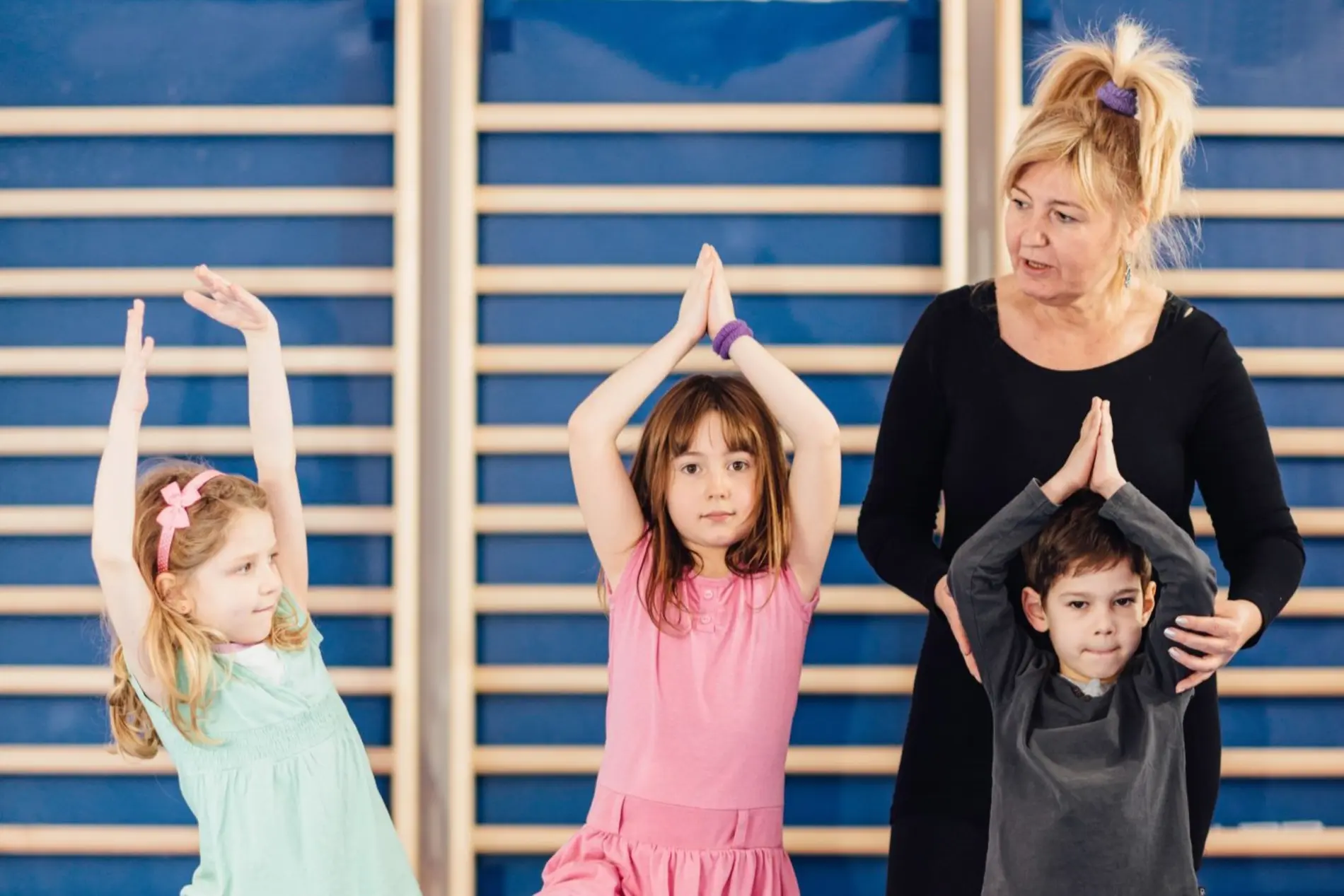 Drei Kinder und eine Lehrerin stehen vor einer Sprossenwand im Turnsaal. Die Kinder halten die Hände gefaltet über den Kopf und nehmen eine Yoga Pose ein