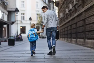 Ein Vater hält seinen Sohn mit Schultasche an der Hand. Sie gehen durch eine verkehrsberuhigte Straße in einer Gemeinde.