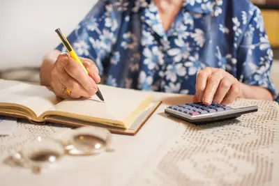 Hände einer älteren Dame bedienen einen Taschenrechner; die rechte Hand schreibt in ein Buch.