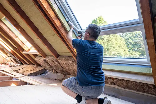 Eine Person arbeitet mit einem Werkzeug an der Abdichtung eines großen Dachfensters in einem ausgebauten Dachraum.