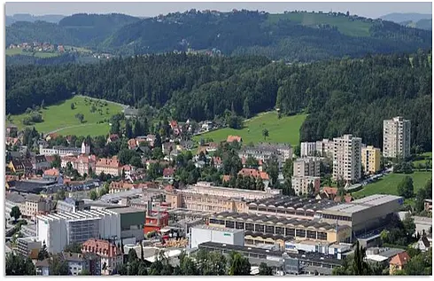 Luftaufnahme eines Industrieareals mit umliegender Stadt und grünen Hügeln im Hintergrund.