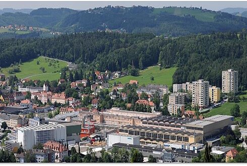 Luftaufnahme eines Industrieareals mit umliegender Stadt und grünen Hügeln im Hintergrund.