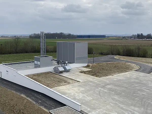 Modernes Heizwerksgebäude mit Lüftungsanlagen und Schornstein, umgeben von Feldern und einer Zufahrtsstraße.