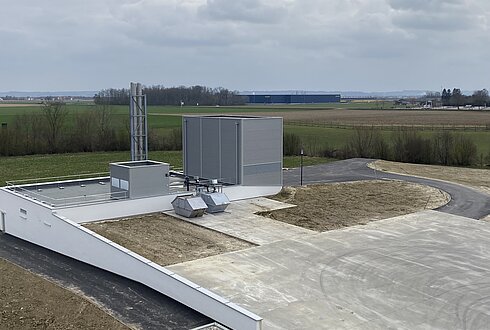 Modernes Heizwerksgebäude mit Lüftungsanlagen und Schornstein, umgeben von Feldern und einer Zufahrtsstraße.