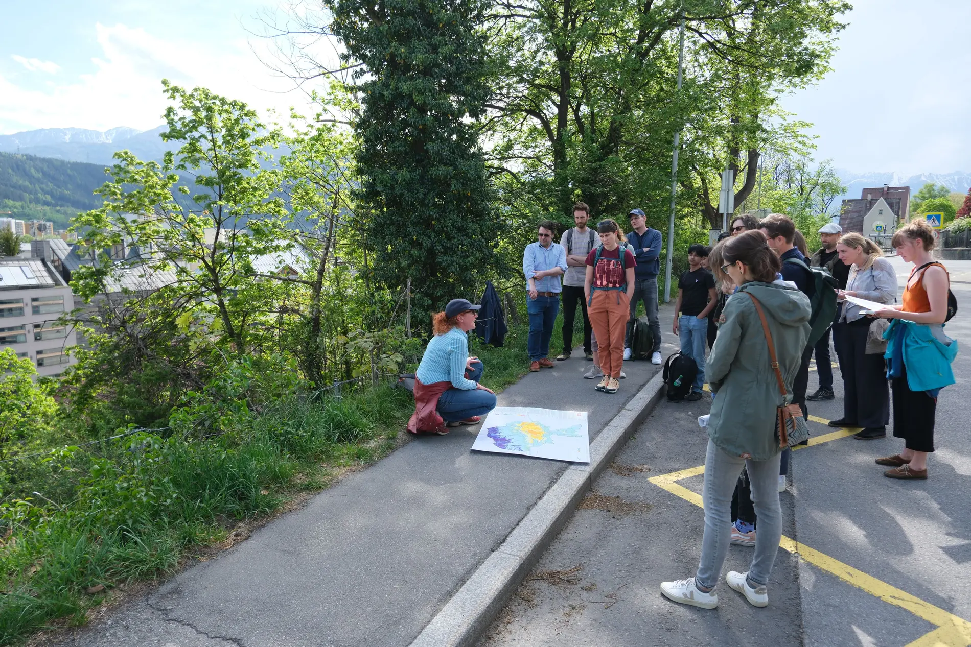 Gruppe von Menschen beim Climate Walk in Innsbruck während des 25. Österreichischen Klimatags 2025. Eine Person kniet und zeigt eine Karte oder ein Poster, die anderen hören zu. Im Hintergrund sind Bäume, Gebäude und Berge zu sehen.